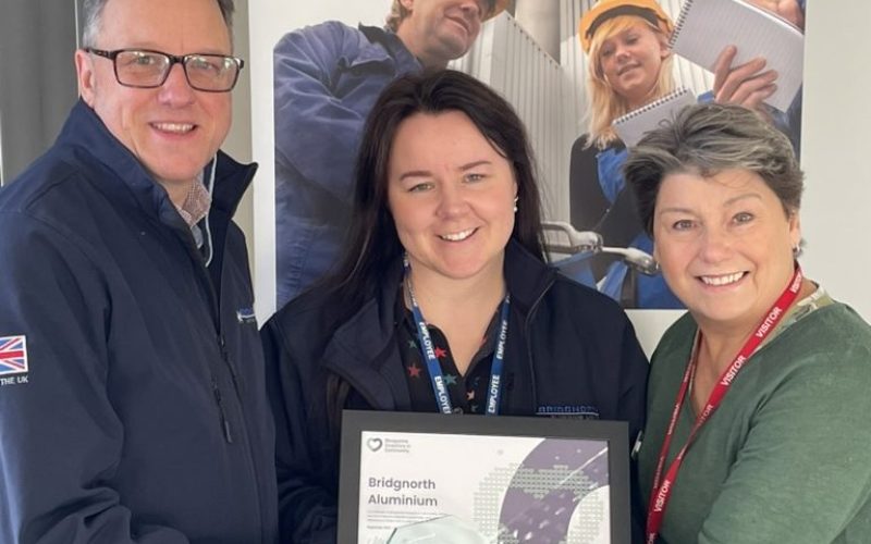 Three people pose together holding a framed certificate or award relating to Bridgnorth Aluminium. A man in a dark jacket and glasses stands on the left, a woman in a dark jacket with a lanyard stands in the centre, and a woman in a green jumper with a red lanyard stands on the right. Behind them is a large display banner showing the Bridgnorth Aluminium company branding and images of workers in orange hard hats at what appears to be an industrial facility.